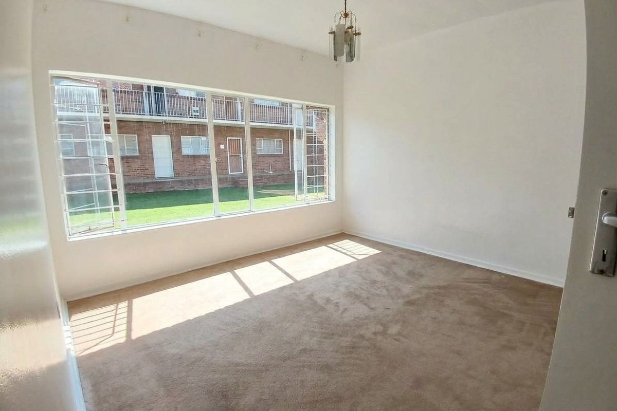 A bright, empty bedroom featuring large windows with security bars, neutral carpeting, and a central ceiling light fixture.