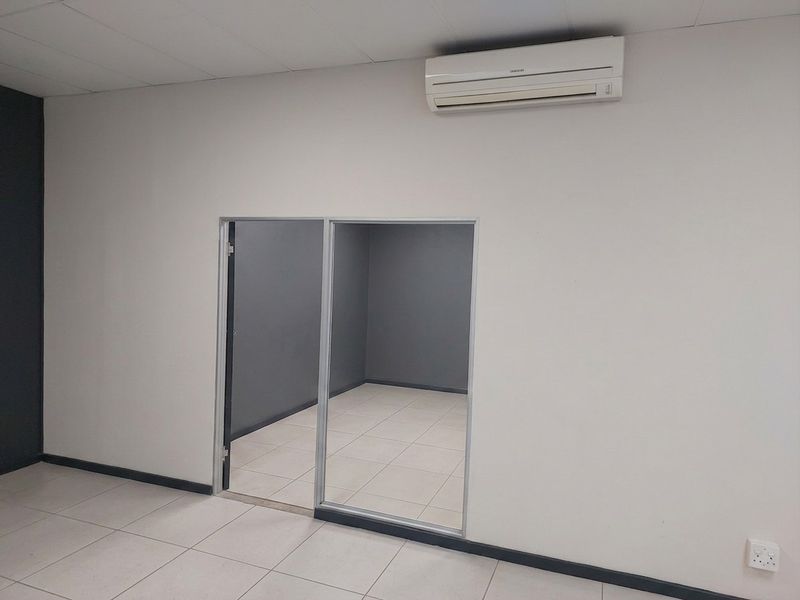 A clean, empty office space featuring tiled flooring, a split-system air conditioner, and a modern aluminum-framed glass partition leading to an adjacent room.