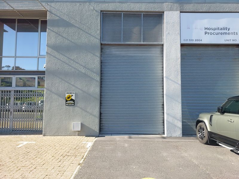 Front exterior view of a commercial office unit featuring a secure roller shutter door, metal security gate, and paved parking area.