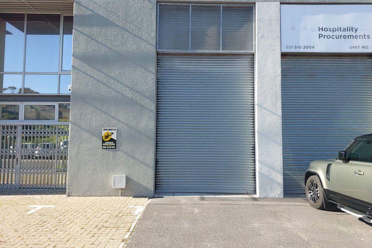 Front exterior view of a commercial office unit featuring a secure roller shutter door, metal security gate, and paved parking area.