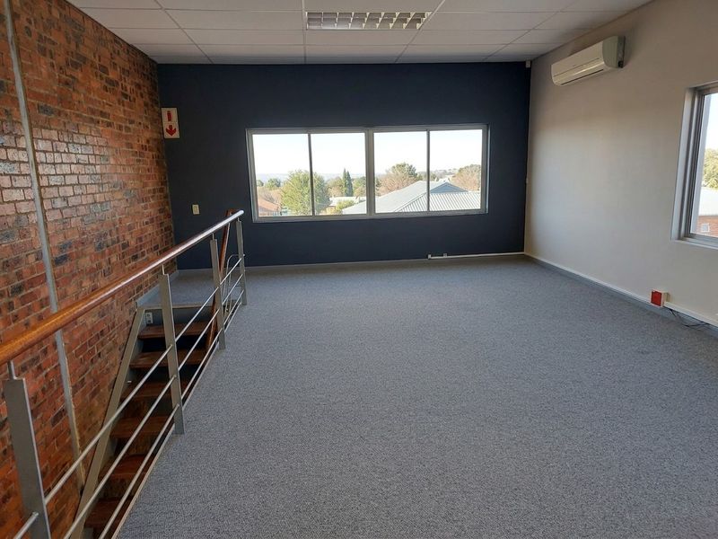 A spacious, empty office area featuring an exposed brick wall, carpeted flooring, a suspended ceiling, and a split-system air conditioner with large windows.