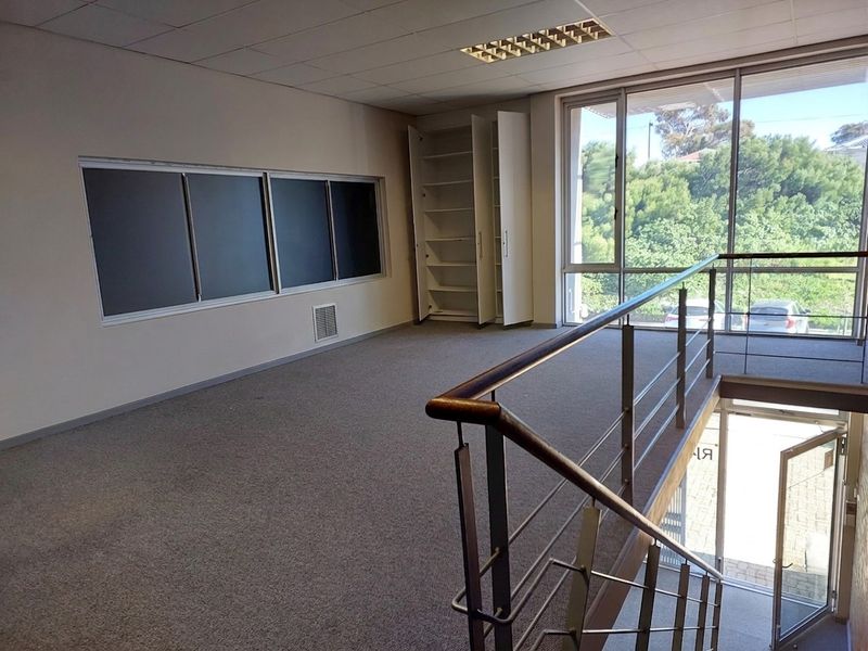 An open-plan office space featuring a suspended ceiling, built-in shelving, carpeted flooring, and large windows overlooking the exterior.