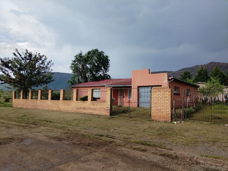 Front view of a single-story house featuring a brick perimeter wall, metal security fencing, and a corrugated metal roof set against a mountain backdrop.