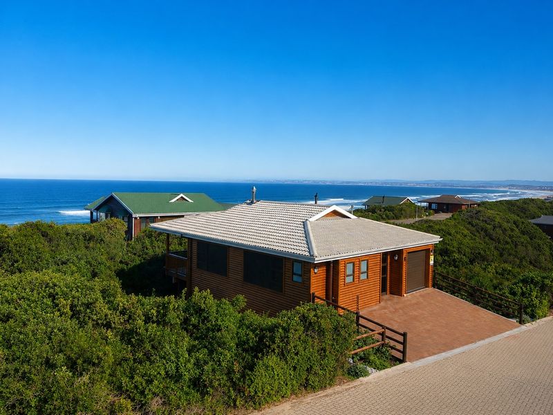 Aerial view of a well-maintained timber log cabin featuring a tiled roof, paved driveway, and scenic ocean views in a coastal setting.