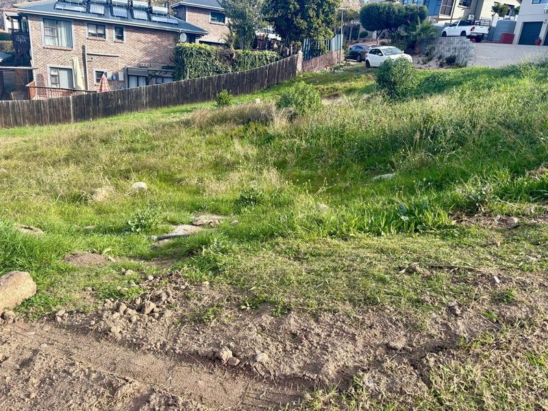 A sloped, undeveloped residential plot featuring natural grass cover and existing boundary fencing in a suburban neighborhood setting.