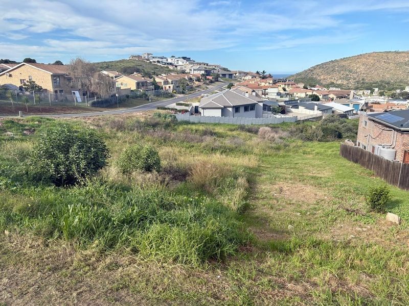A vacant residential plot featuring sloped terrain and natural grass, situated in an established neighborhood with surrounding residential properties.
