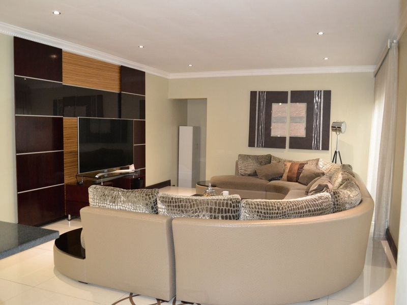 A spacious living room featuring a modern built-in media wall unit, recessed downlighting, crown molding, and polished tiled flooring.