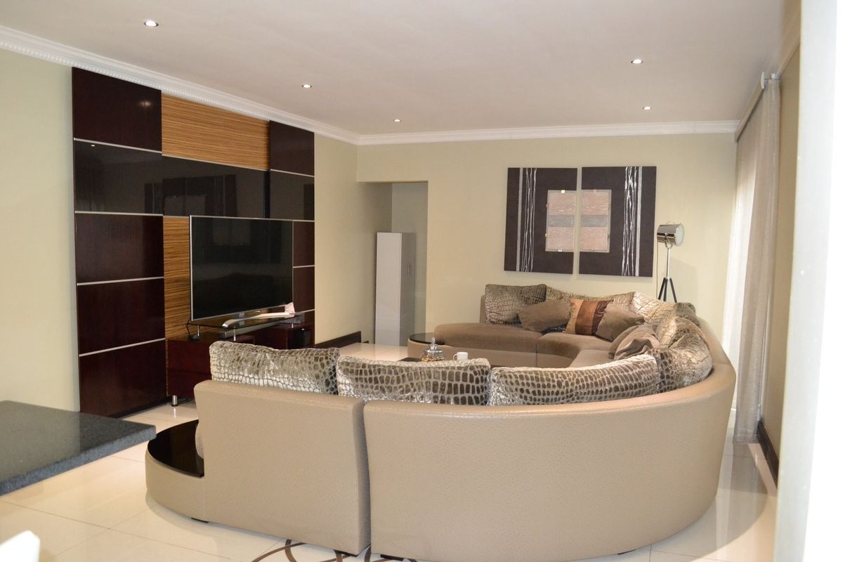 A spacious living room featuring a modern built-in media wall unit, recessed downlighting, crown molding, and polished tiled flooring.
