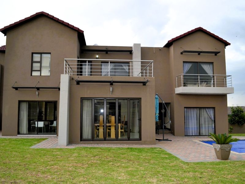 Rear view of a double-story residential property featuring a paved patio, swimming pool, landscaped garden, and upper-level balconies with stainless steel railings.