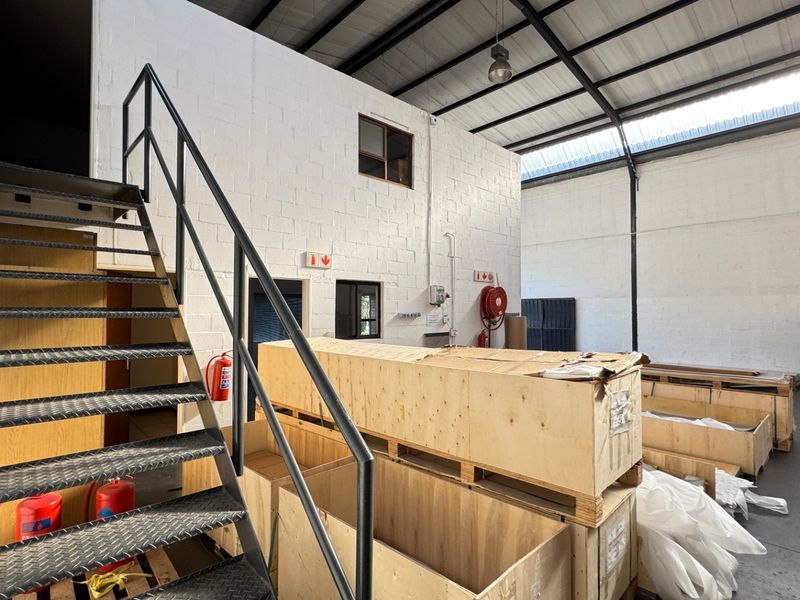 Industrial warehouse space featuring high ceilings with exposed steel trusses, concrete flooring, and a metal staircase leading to an upper level.