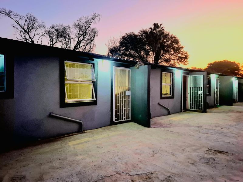 Rear exterior view of a residential building featuring multiple entry points with security gates, casement windows, and a concrete paved courtyard area at dusk.