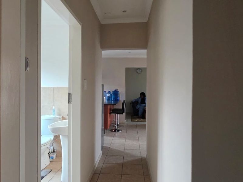 A view through a hallway connecting to a bathroom and an open-plan living area with tiled flooring and neutral wall finishes.