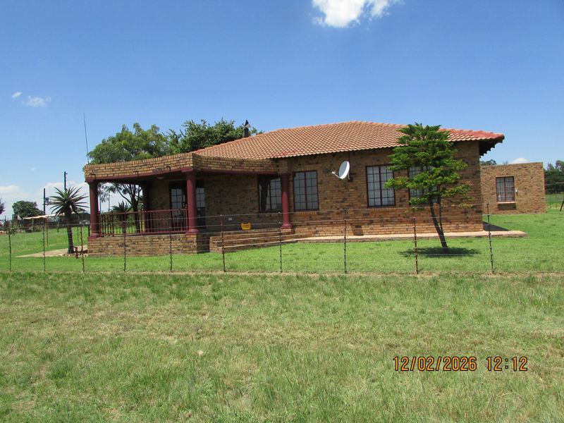 A well-maintained face brick farmhouse featuring a covered patio, tiled roof, and surrounding lawn area on a rural property.