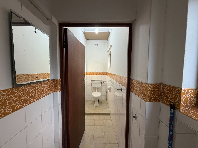 A dated bathroom featuring a toilet, tiled walls with decorative borders, and tiled flooring, requiring cosmetic updates and repairs to the ceiling and mirror.