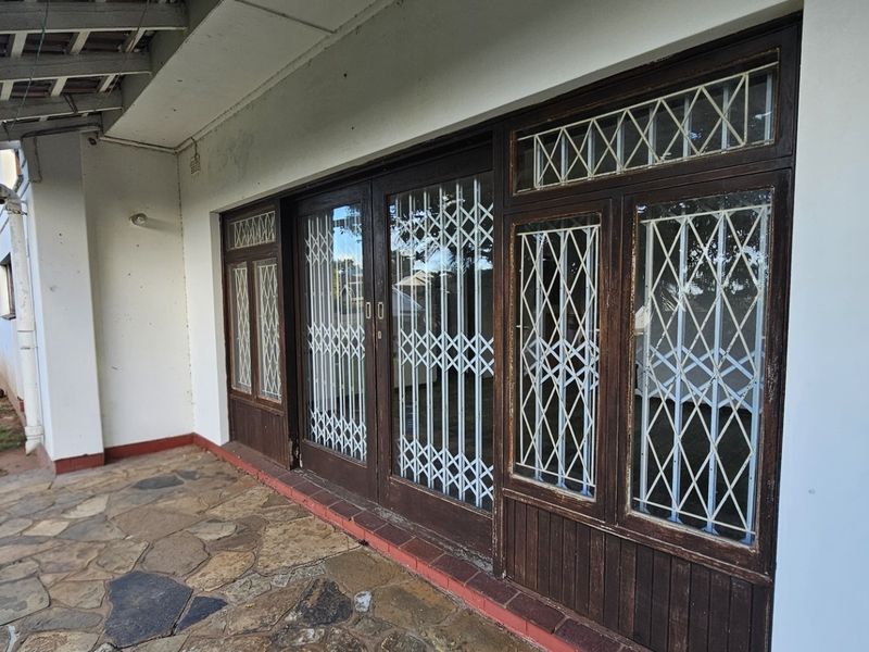 A covered patio area featuring stone paving and timber-framed glass doors with security gates, showing signs of wear and requiring maintenance.