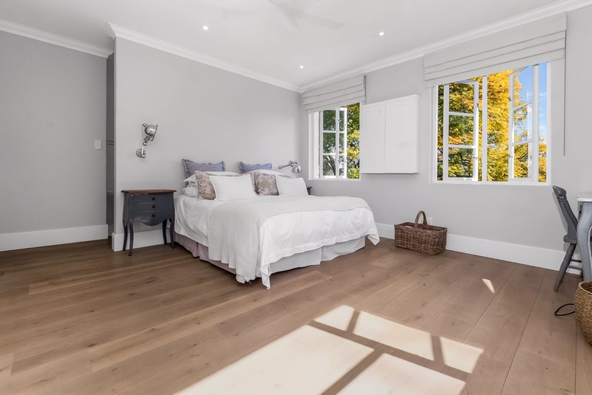 Spacious bedroom featuring light-toned engineered timber flooring, recessed downlighting, crown molding, and large casement windows providing natural light and garden views.