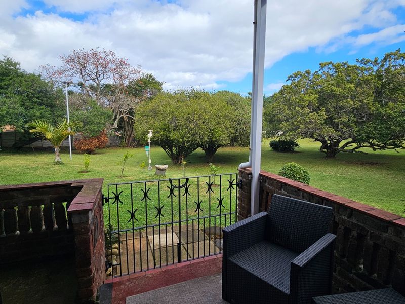 A covered patio area featuring a brick balustrade and metal security gate, overlooking a spacious, well-maintained garden with mature trees.