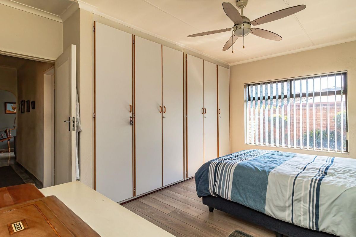 A functional bedroom featuring extensive built-in cupboards, a ceiling fan, and vertical blinds, finished with modern laminate flooring.