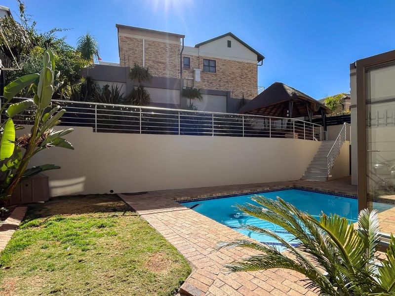 A well-maintained outdoor pool area featuring brick paving, a retaining wall with stainless steel balustrades, and a thatched lapa for outdoor entertainment.