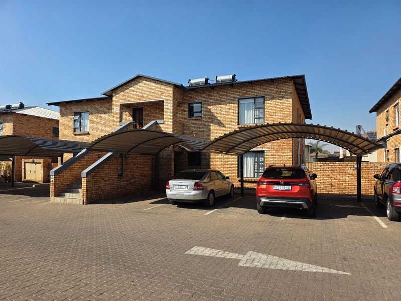 Exterior view of a face-brick apartment complex featuring covered parking carports, paved driveway, and solar water heating systems on the roof.