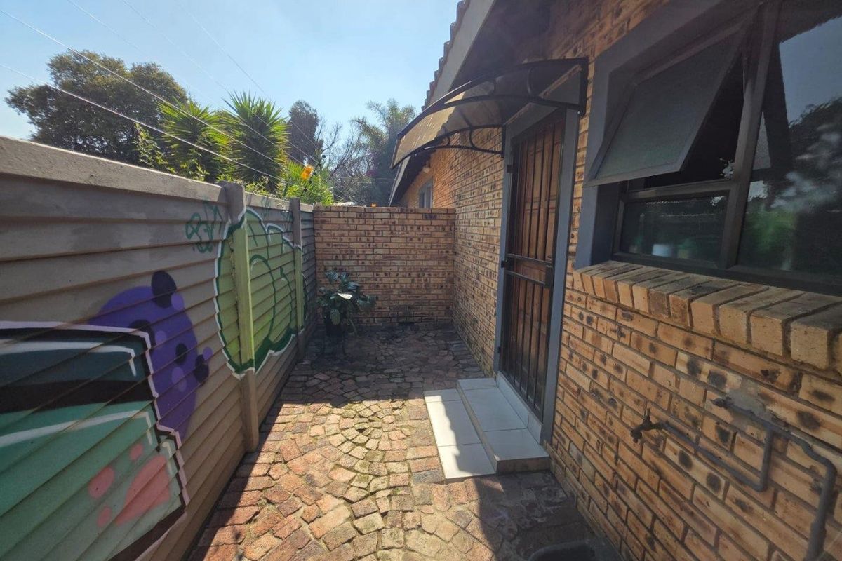 A rear exterior view showing a brick facade, paved walkway, security gate, and a boundary wall with graffiti.