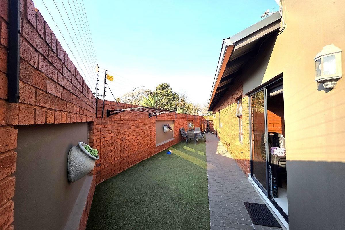 A side exterior view featuring a paved walkway, artificial grass lawn, and a high brick boundary wall equipped with electric fencing.