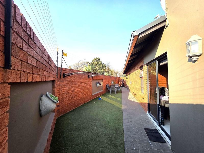 A side exterior view featuring a paved walkway, artificial grass lawn, and a high brick boundary wall equipped with electric fencing.