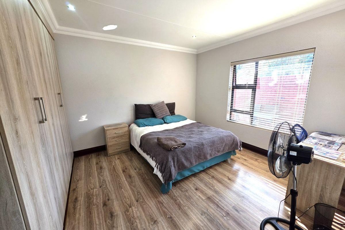 A well-maintained bedroom featuring modern laminate flooring, extensive built-in cupboards, recessed ceiling lighting, and large windows with fitted venetian blinds.