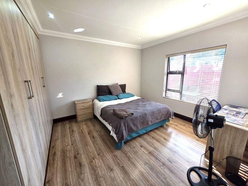 A well-maintained bedroom featuring modern laminate flooring, extensive built-in cupboards, recessed ceiling lighting, and large windows with fitted venetian blinds.