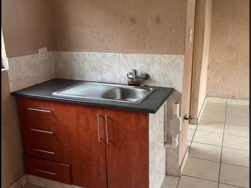 A compact kitchen area featuring a single sink unit with built-in cabinetry, tiled flooring, and a tiled splashback, requiring cosmetic updates.