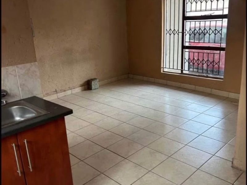 An empty kitchen area featuring tiled flooring, basic cabinetry with a stainless steel sink, and a window fitted with security bars.