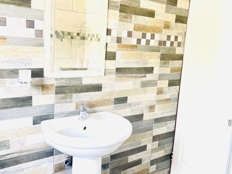 A clean bathroom featuring a white pedestal basin, wall-mounted mirror, and decorative multi-toned wall tiling.