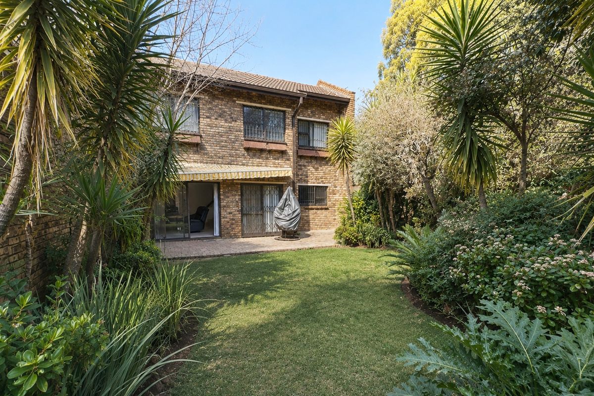 Rear view of a two-story brick townhouse featuring a paved patio, retractable awning, and a well-established private garden.