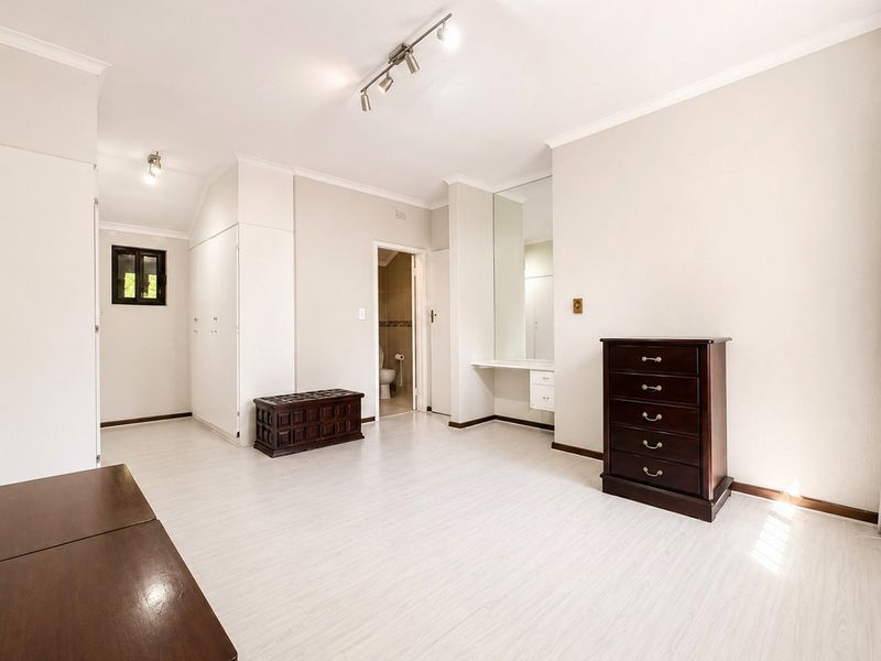 Spacious bedroom featuring light laminate flooring, built-in cupboards, a dedicated vanity area with mirror, and modern track lighting.