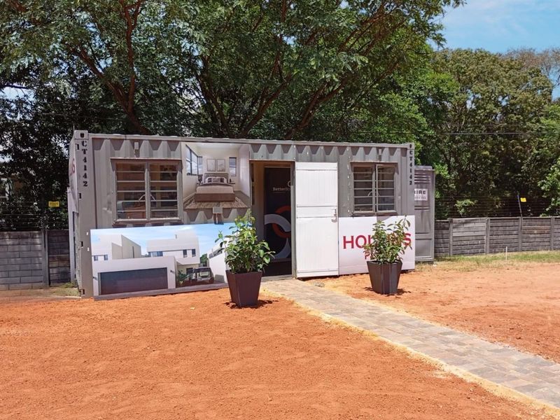 A temporary sales office or site container structure located on an undeveloped plot with paved access and security fencing.