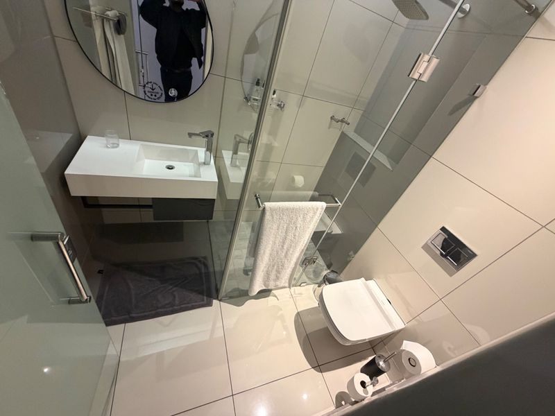 Modern bathroom featuring a wall-mounted vanity, glass shower enclosure, and wall-mounted toilet with neutral floor-to-ceiling tiling.