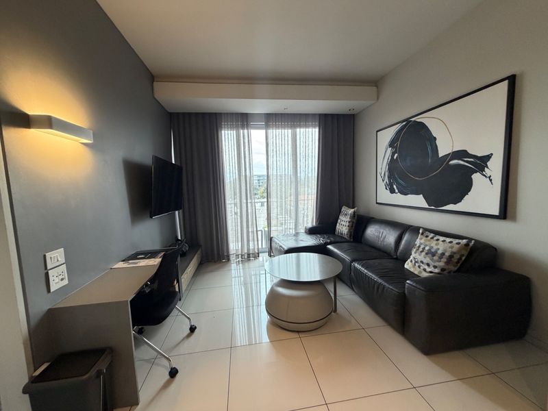 A modern living room featuring tiled floors, a built-in desk, and a sliding glass door leading to a balcony, creating a bright and functional space.