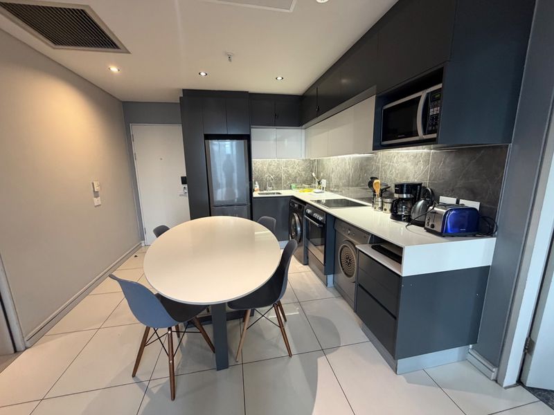 Modern open-plan kitchen featuring sleek dark cabinetry, stone countertops, integrated appliances, and tiled flooring, seamlessly flowing into the dining area.