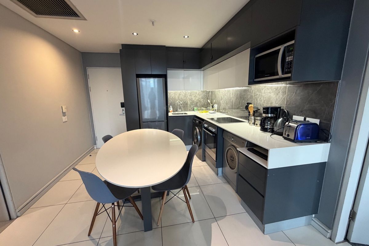 Modern open-plan kitchen featuring sleek dark cabinetry, stone countertops, integrated appliances, and tiled flooring, seamlessly flowing into the dining area.