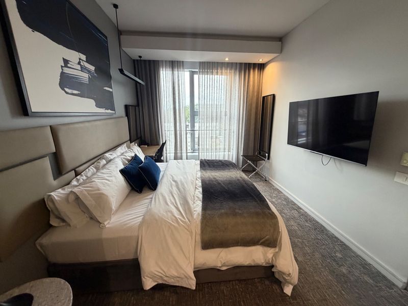 A modern, well-maintained bedroom featuring an upholstered headboard, recessed ceiling lighting, and a wall-mounted television with access to a balcony.