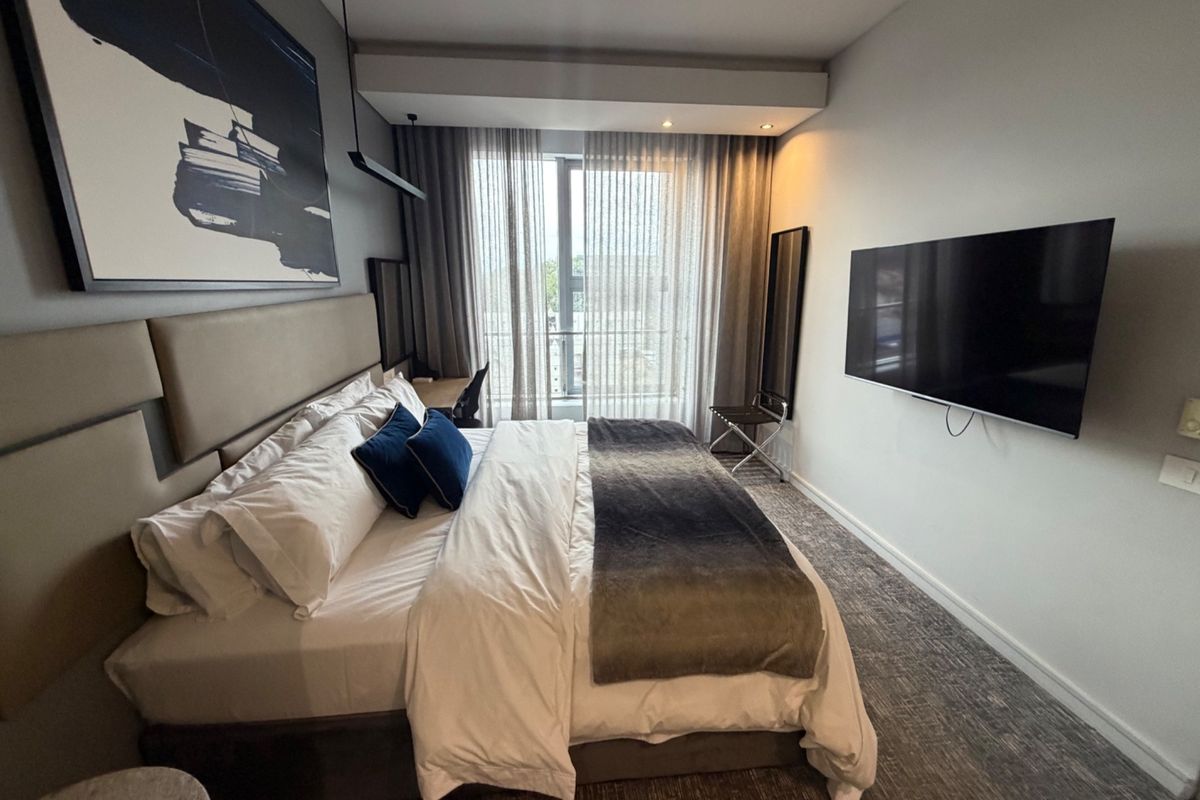 A modern, well-maintained bedroom featuring an upholstered headboard, recessed ceiling lighting, and a wall-mounted television with access to a balcony.