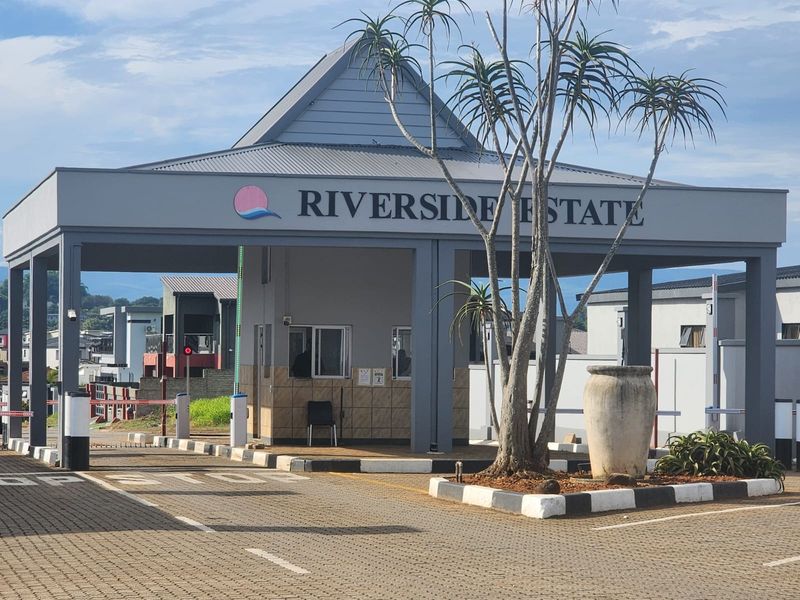 The main entrance gatehouse for Riverside Estate, featuring a paved driveway, security boom gates, and a well-maintained structure.