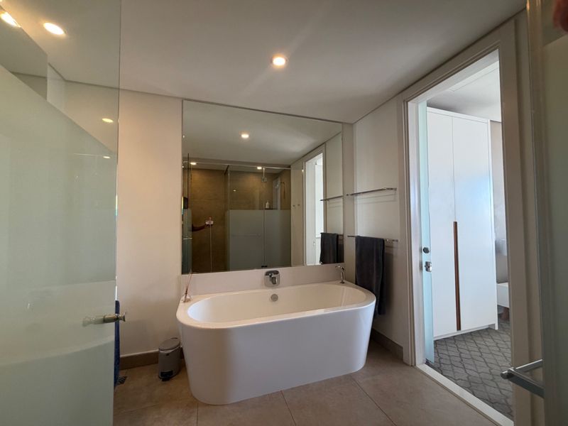 Modern bathroom featuring a sleek freestanding bathtub, large wall-mounted mirror, recessed lighting, and a glass-enclosed shower area with neutral floor tiling.