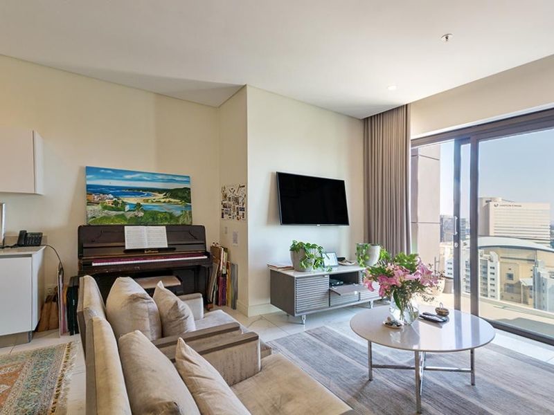 A bright and modern living room featuring tiled floors, recessed lighting, and large sliding glass doors that open to a balcony with city views.