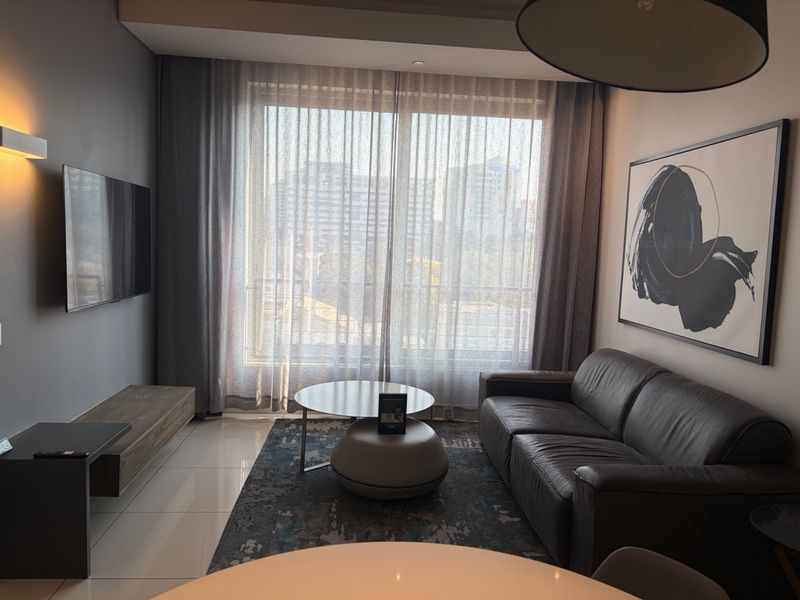 A modern, well-lit living room featuring tiled floors, neutral wall tones, and a large window providing natural light and urban views.