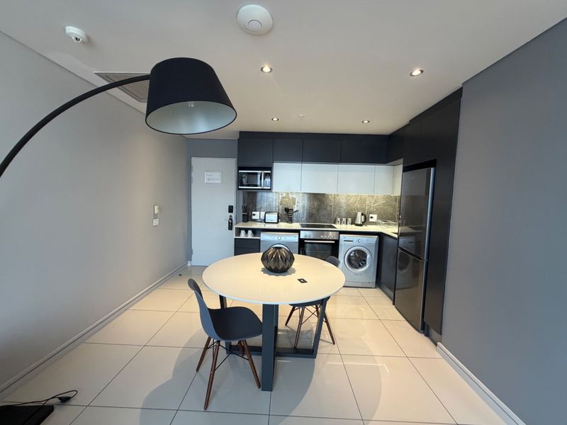 Modern open-plan kitchen featuring sleek dark and white cabinetry, stone-look backsplash, integrated appliances, and tiled flooring, seamlessly flowing into the dining area.