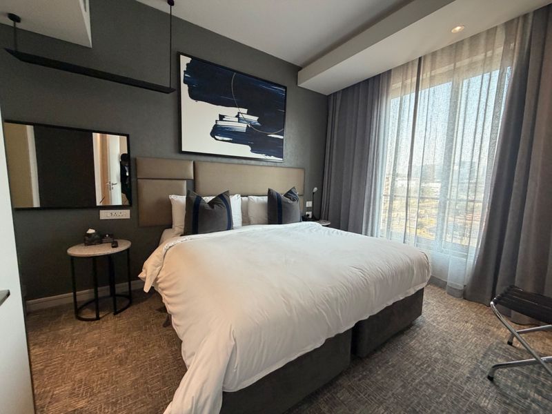 A modern, well-appointed bedroom featuring a neutral color palette, integrated lighting, and large windows with floor-to-ceiling curtains.