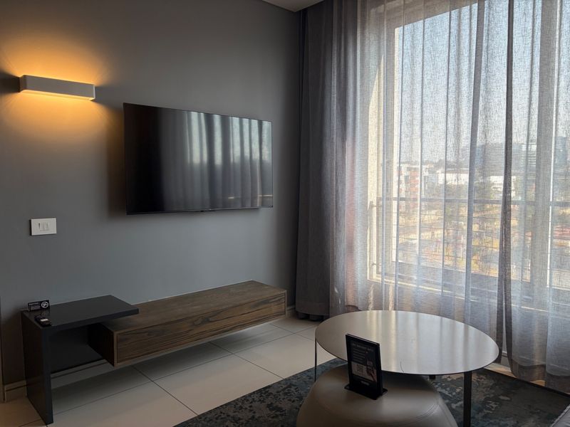 Modern living room featuring a wall-mounted television, floating media console, sleek tiled flooring, and large windows with sheer curtains providing ample natural light.