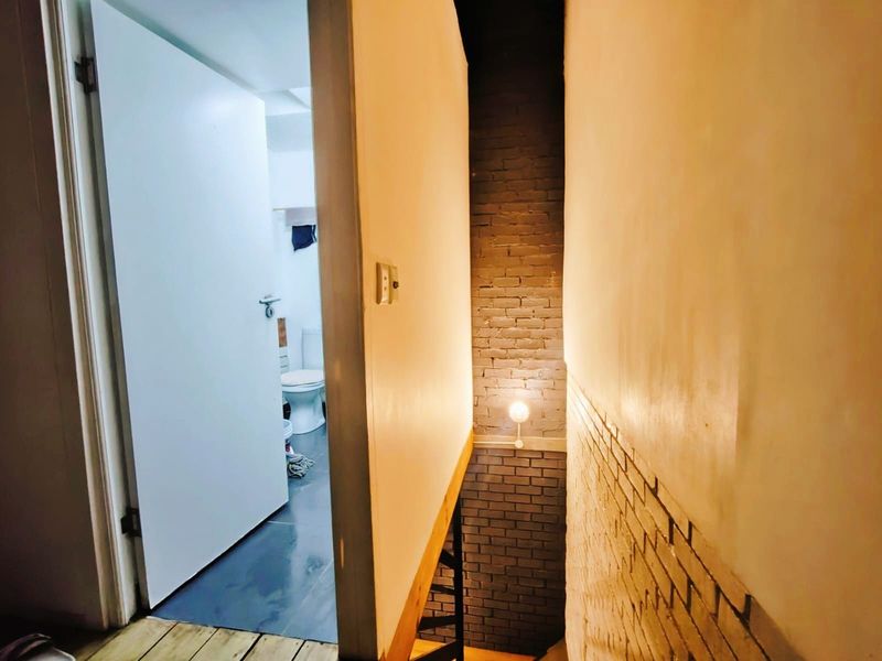 A narrow hallway featuring exposed brickwork and a staircase, leading to an adjacent bathroom area with visible wear on the wall finishes.