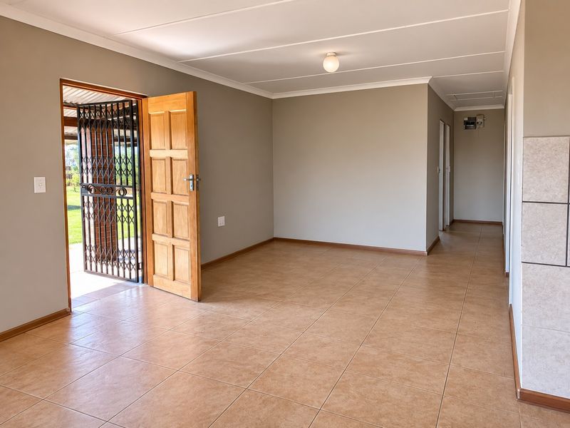A bright, empty living area featuring neutral wall tones, tiled flooring, and direct access to the exterior through a timber door with a security gate.
